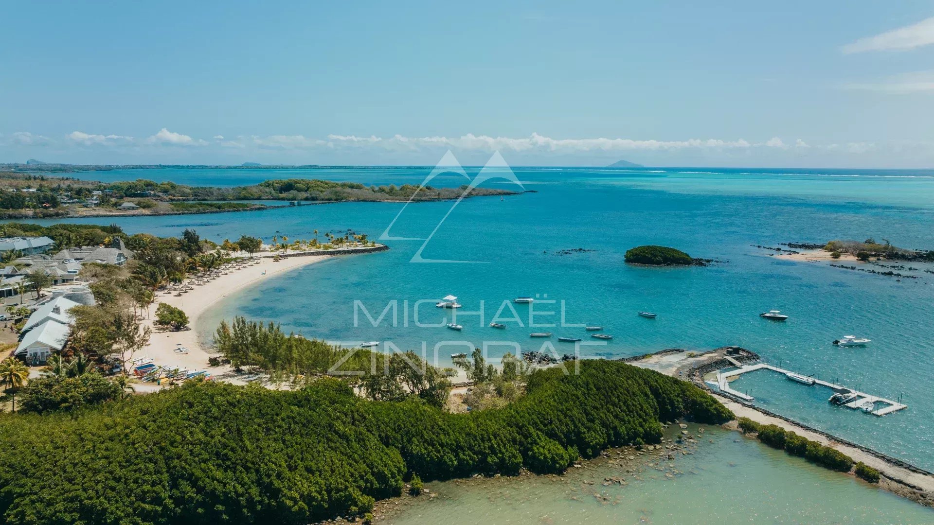 Aerial view of a tropical coastline with a curved white-sand beach, palm trees, and turquoise water with several boats.