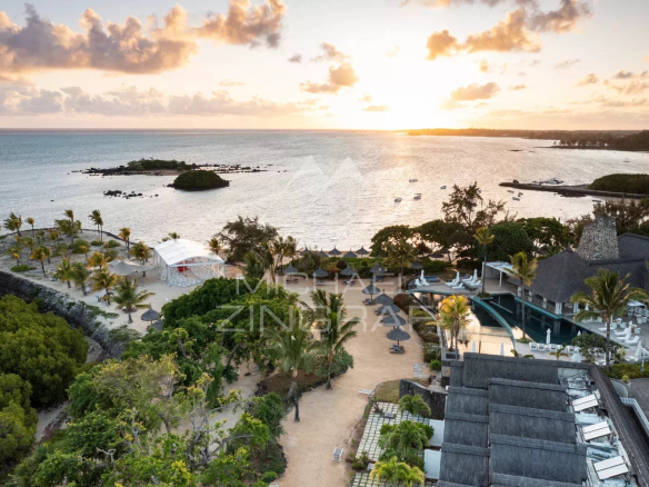 Sunset over a tropical resort: sandy beach, palm trees, and a pool area with lounge chairs by the sea.