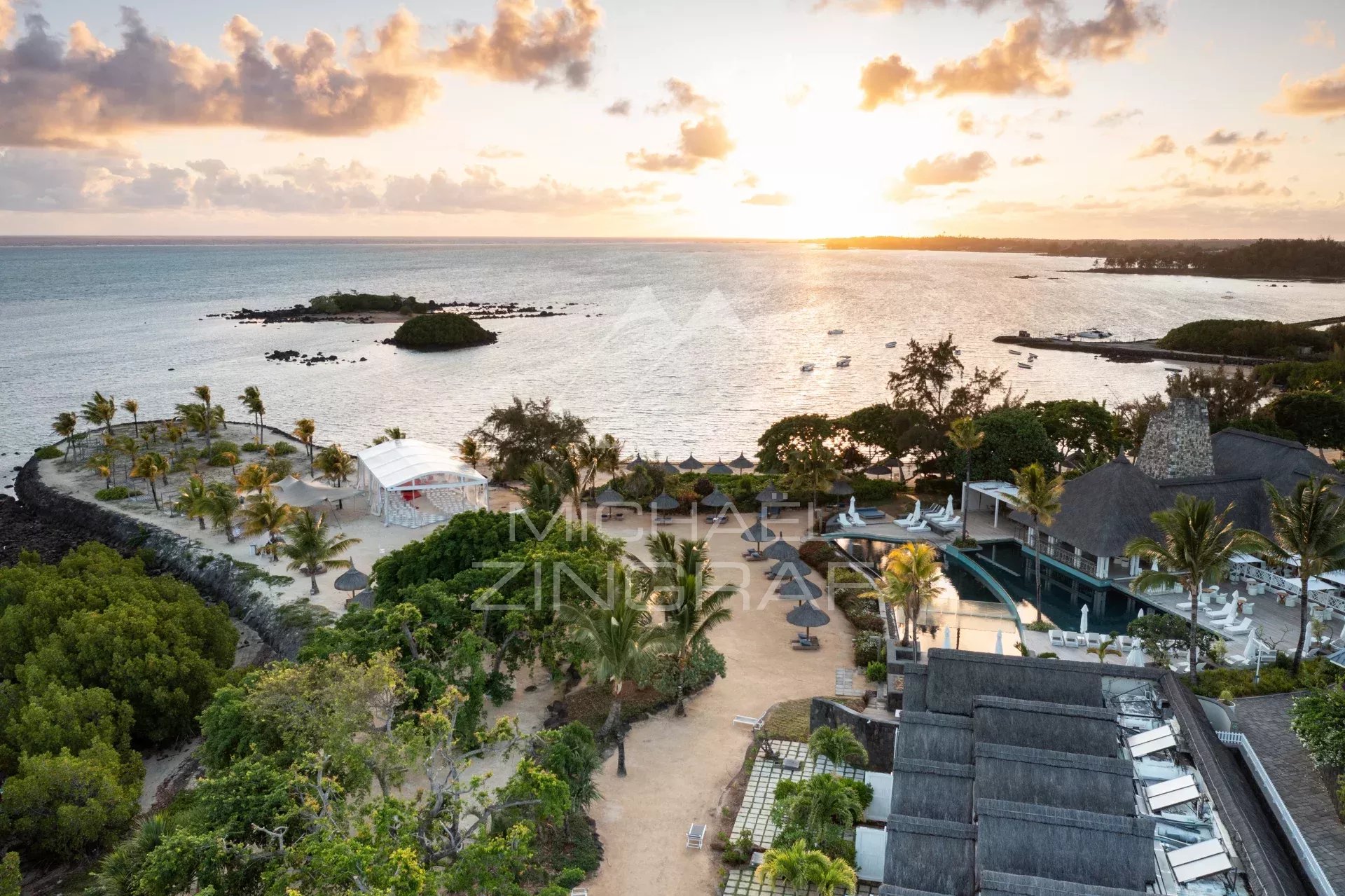 Sunset over a tropical resort: sandy beach, palm trees, and a pool area with lounge chairs by the sea.