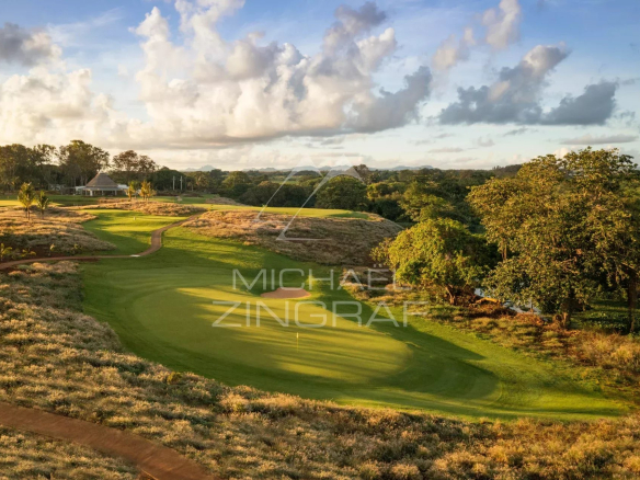 A sunlit golf course with a green, sand bunker, and winding path, bordered by shrubs and trees, with a clubhouse in the distance.