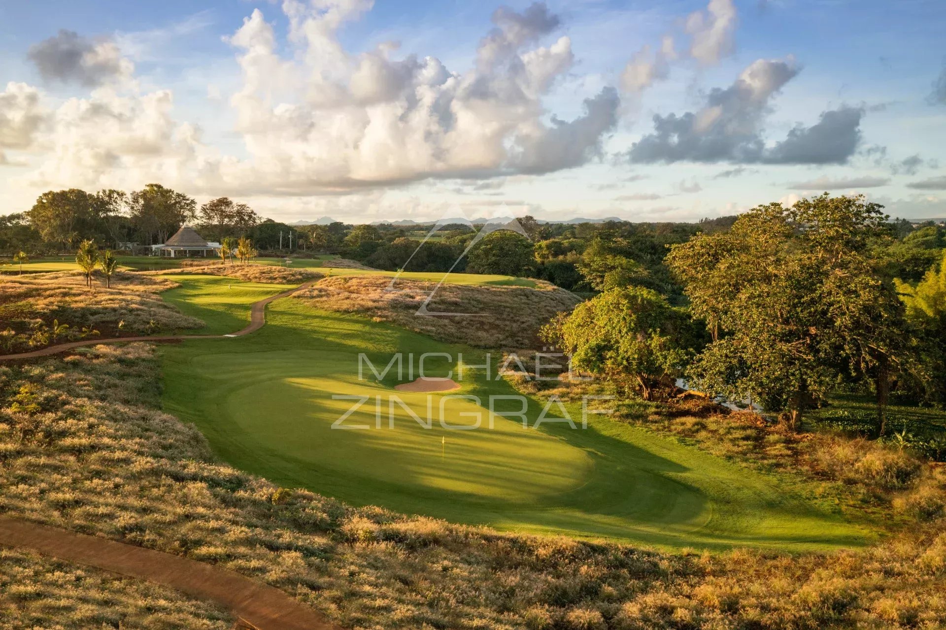A sunlit golf course with a green, sand bunker, and winding path, bordered by shrubs and trees, with a clubhouse in the distance.