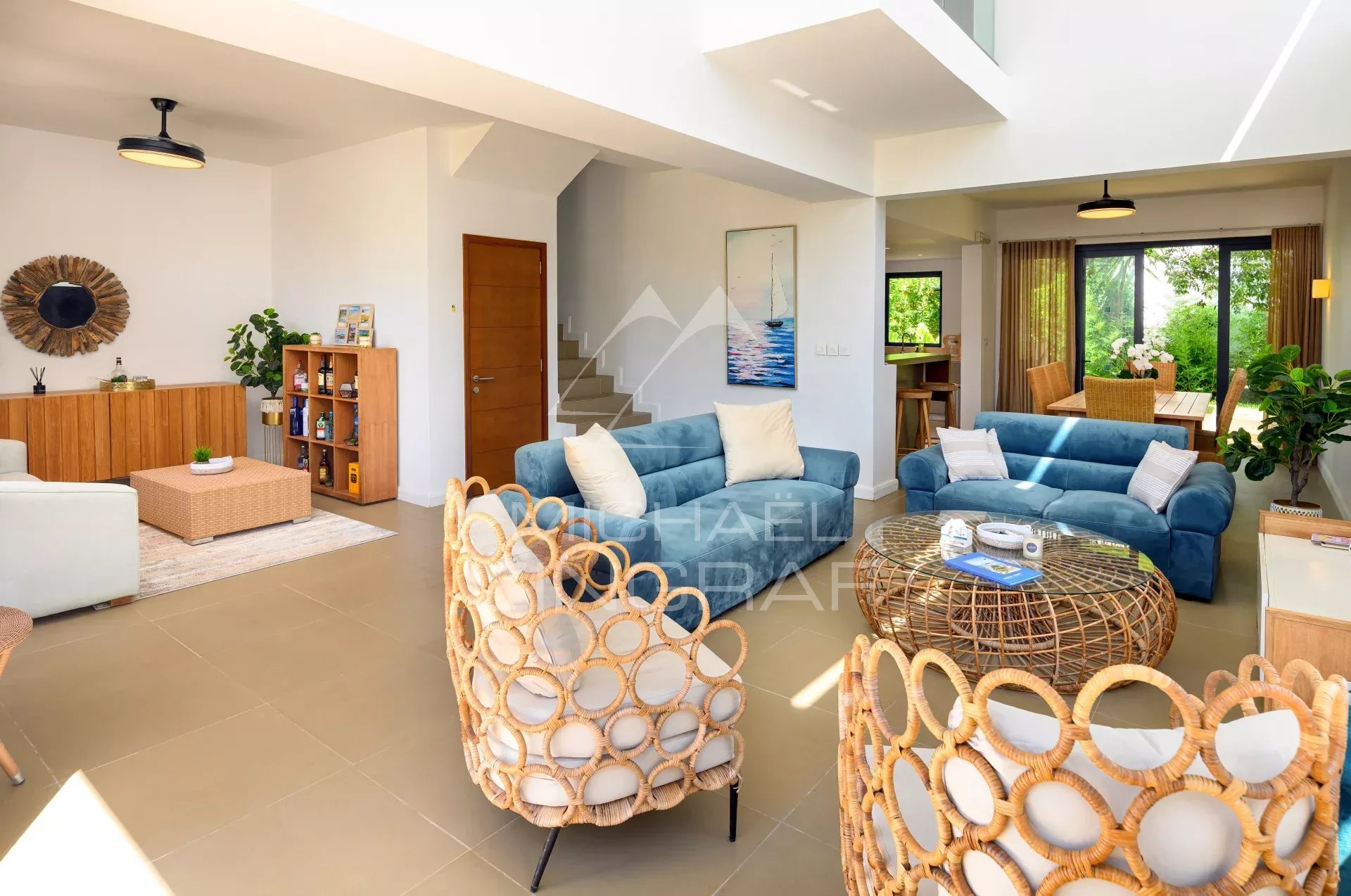 Bright open-concept living room with blue sofas, wicker chairs, and a round wicker coffee table in a sunlit space.
