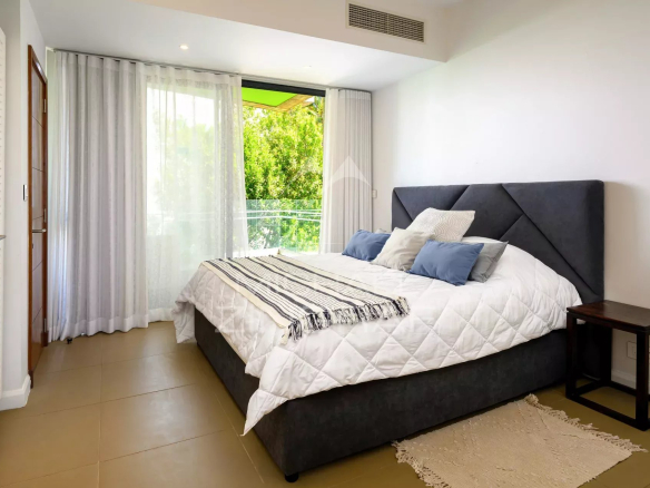 Bedroom with a large dark gray upholstered bed, white quilt, and blue pillows; sliding glass door with sheer curtains to a green outdoor view.