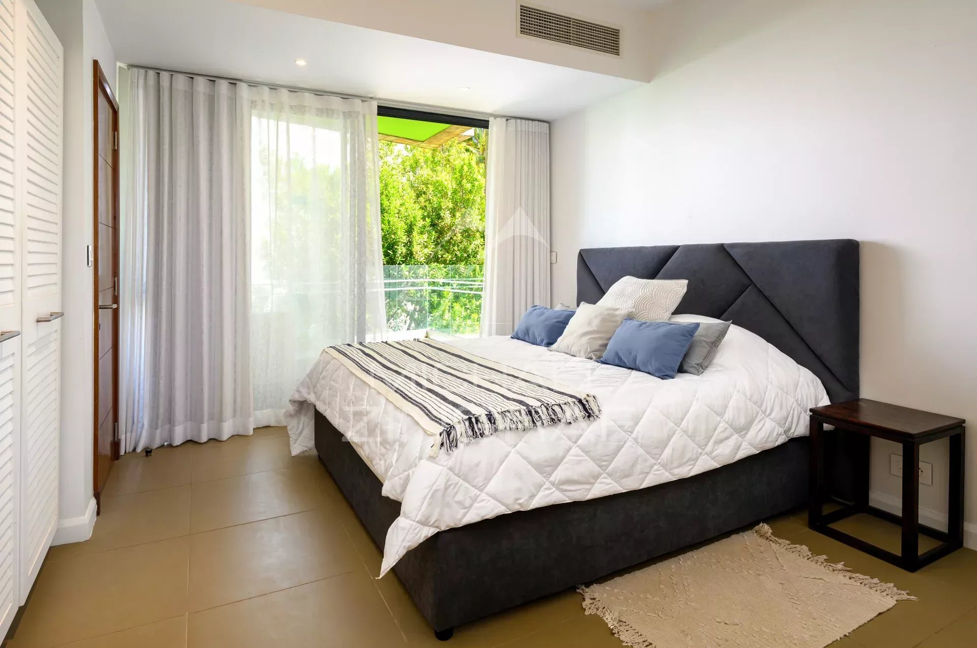 Bedroom with a large dark gray upholstered bed, white quilt, and blue pillows; sliding glass door with sheer curtains to a green outdoor view.