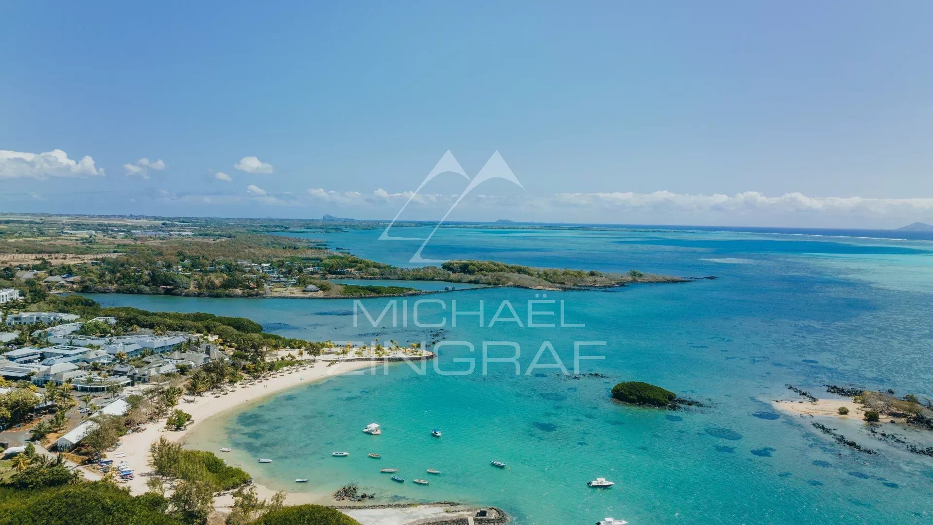 Aerial view of a tropical coastline with a white-sand beach, turquoise water, and boats anchored near shore under a clear blue sky.