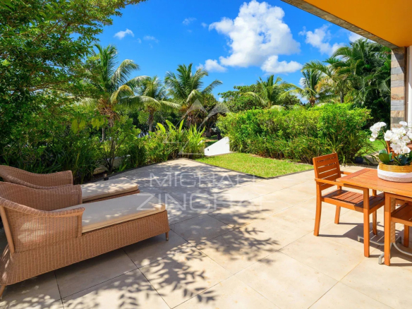 Sunny tropical patio with wicker lounge chairs on the left and a wooden dining table with chairs on the right, surrounded by lush greenery.