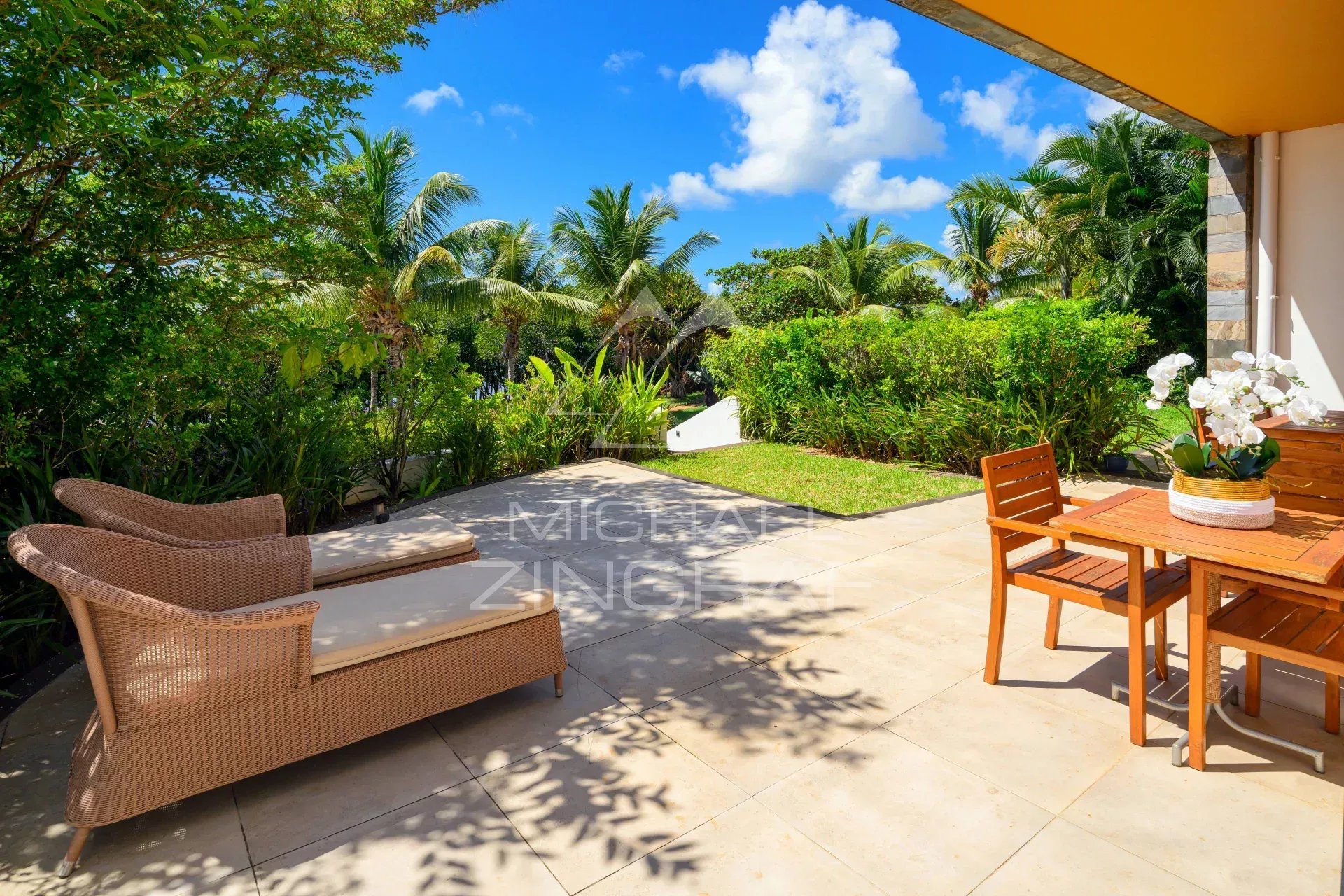Sunny tropical patio with wicker lounge chairs on the left and a wooden dining table with chairs on the right, surrounded by lush greenery.