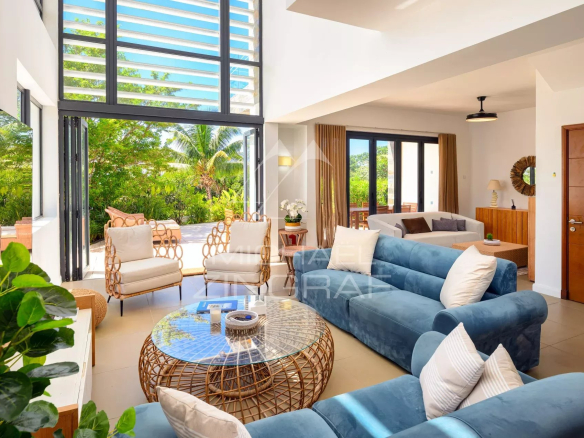 Bright living room with blue sofas and a round wicker coffee table, open glass doors to a sunny patio with tropical plants outside.
