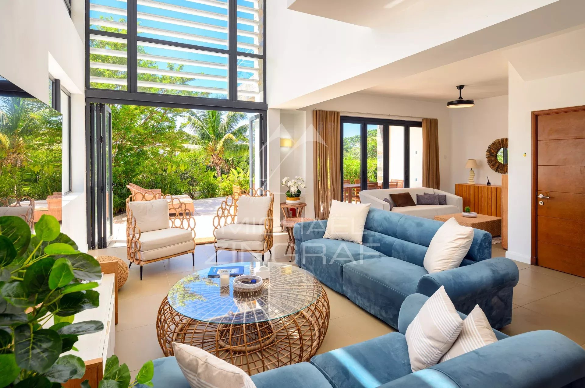 Bright living room with blue sofas and a round wicker coffee table, open glass doors to a sunny patio with tropical plants outside.