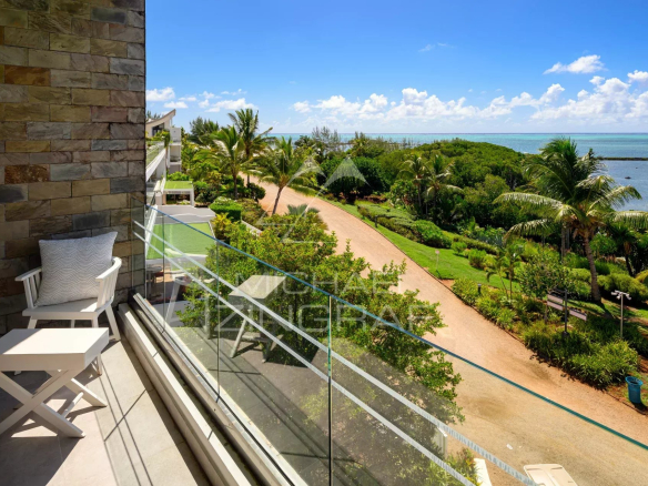 Balcony with a white chair and small table, glass railing, and a stone wall, overlooking a tropical garden path toward the ocean.