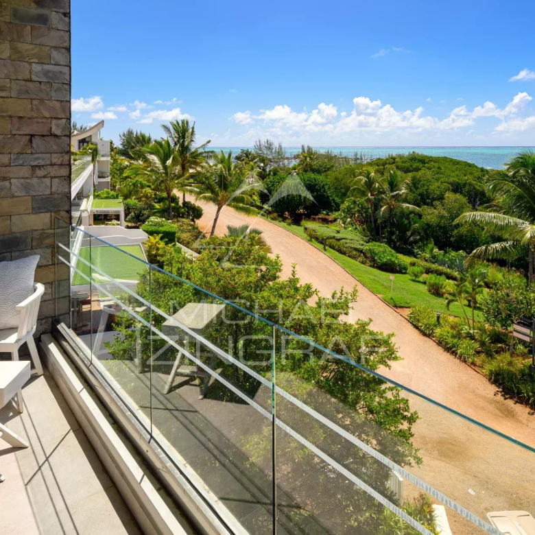 Balcony with a white chair and small table, glass railing, and a stone wall, overlooking a tropical garden path toward the ocean.