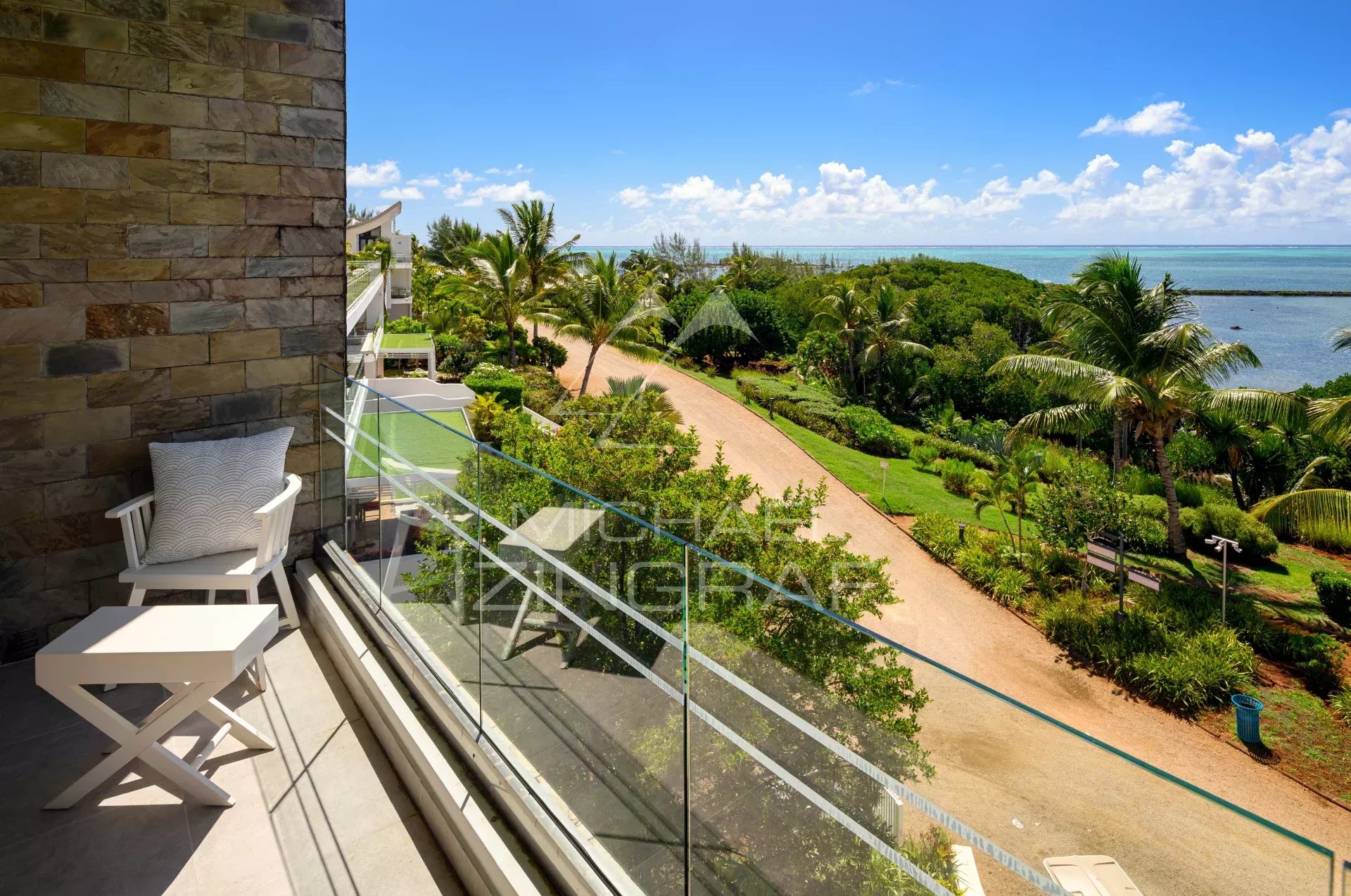 Balcony with a white chair and small table, glass railing, and a stone wall, overlooking a tropical garden path toward the ocean.