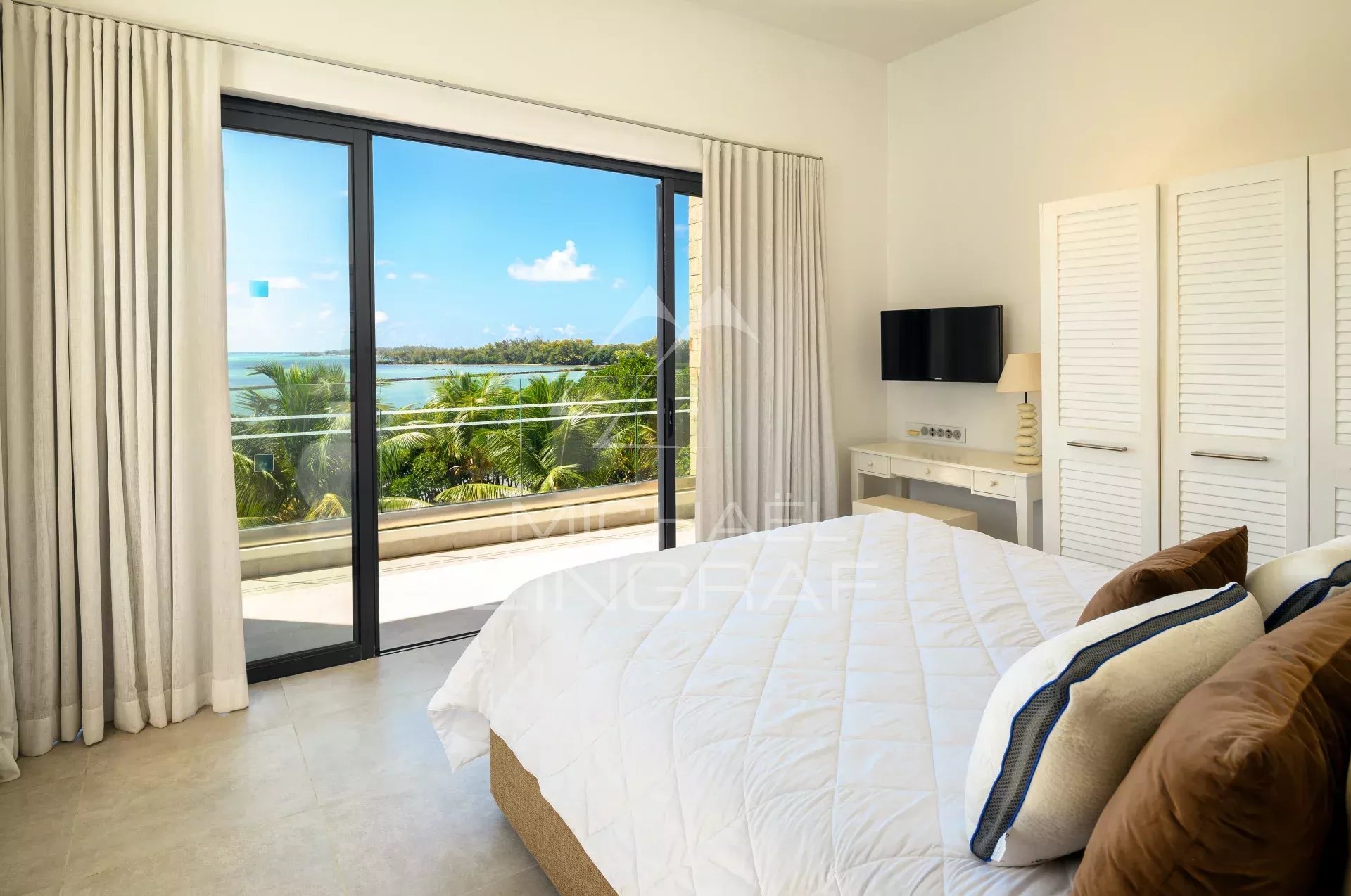 Bedroom with a white quilted bed facing a sliding glass door to a balcony and tropical ocean view.