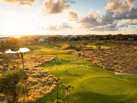 Sunset over a green golf course with patches of dry brush, water hazard, and distant houses on the horizon