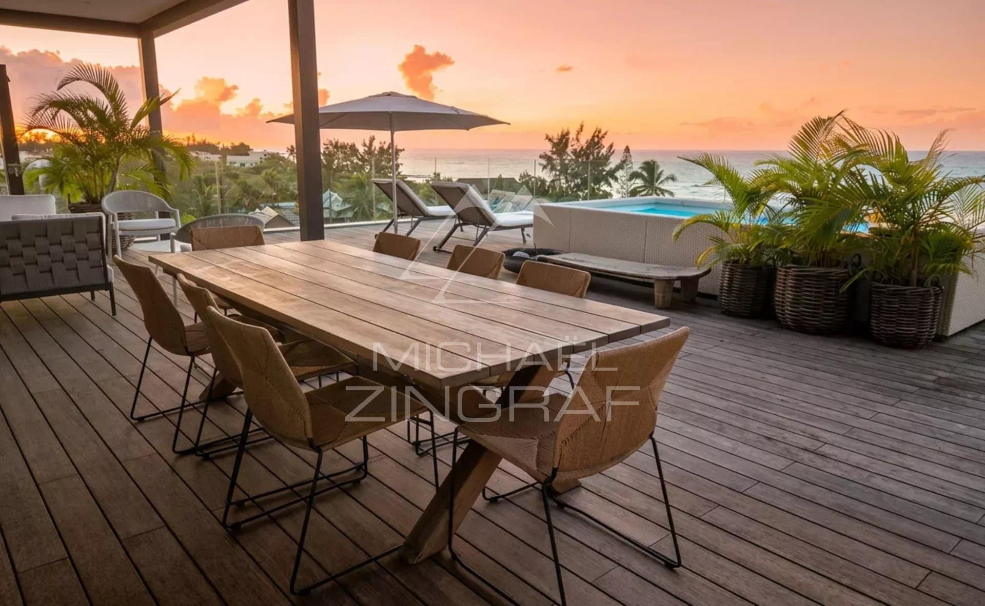 Terrasse d'un penthouse avec vue sur la mer au coucher de soleil, mobilier moderne en bois et chaises en métal.