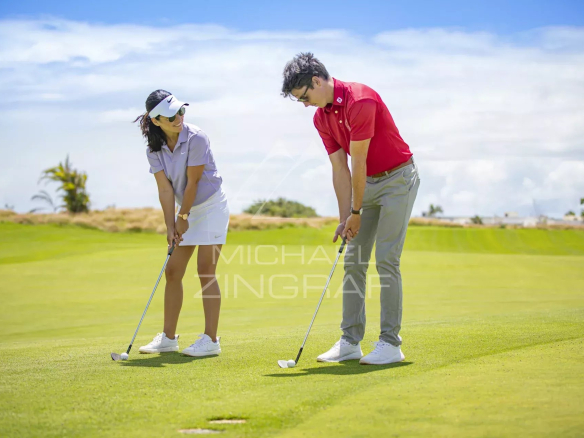 Deux golfeurs s'entraînent sur un green, avec un ciel bleu et des collines en arrière-plan.