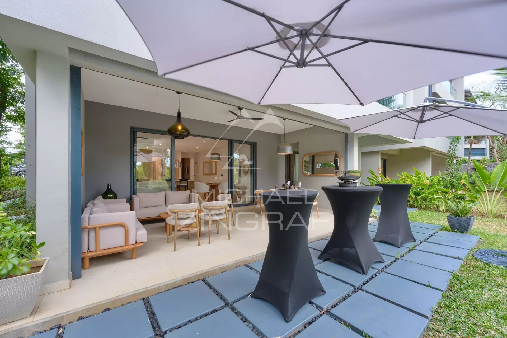 Apartment terrace with modern furniture and parasols, surrounded by greenery.