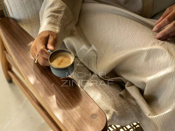 Personne assise sur un fauteuil en bois, tenant une tasse de café dans un intérieur lumineux.