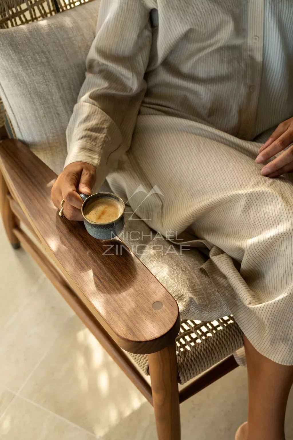 Détente avec vue Personne assise sur un fauteuil en bois, tenant une tasse de café dans un intérieur lumineux.