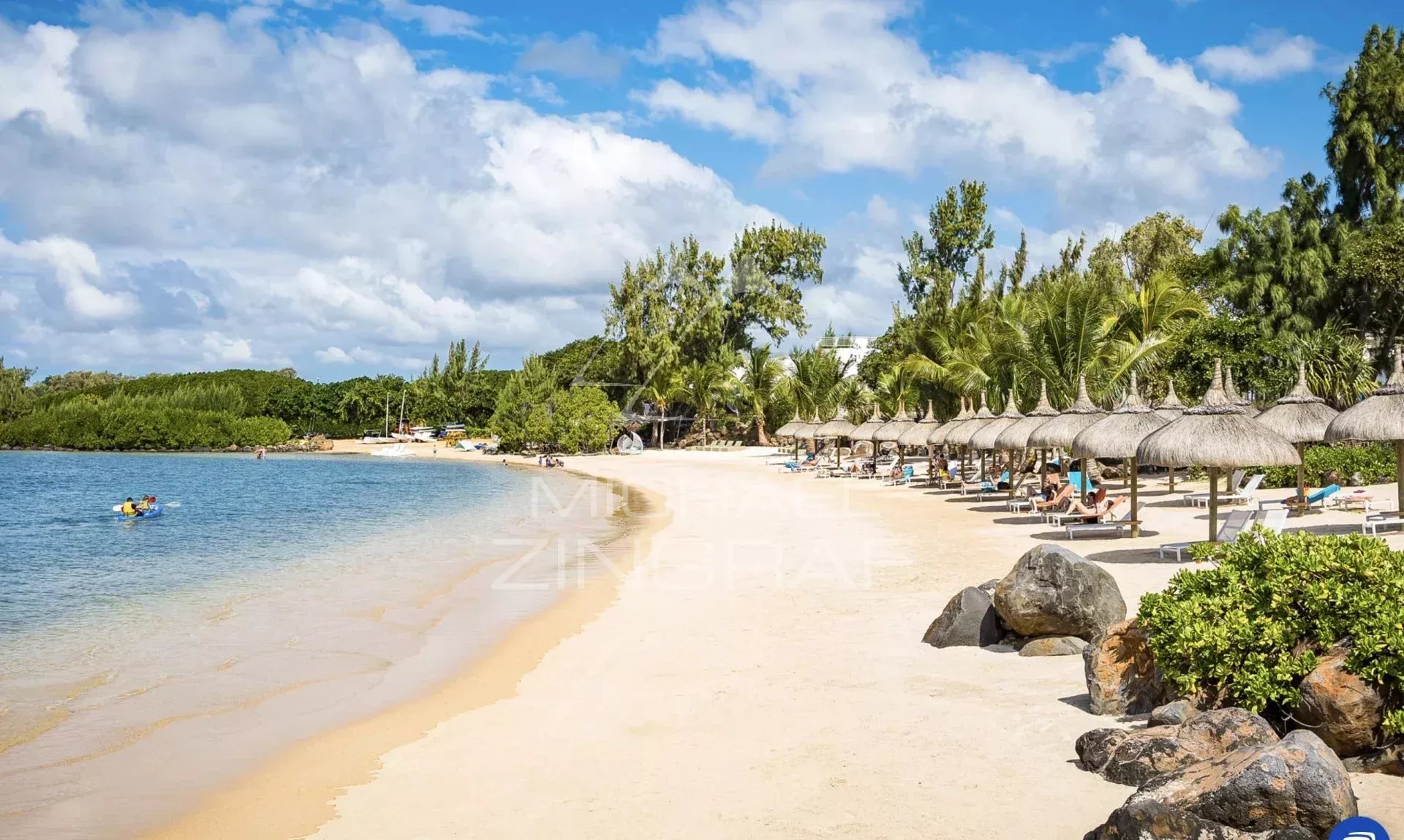 Plage de sable avec parasols et chaises longues, bordée par des eaux calmes et des palmiers.