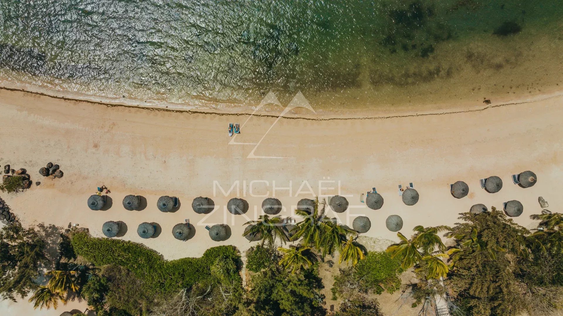 Vue aérienne d'une plage avec parasols et palmiers, près d'une mer calme.