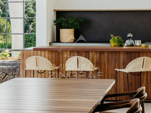 Intérieur moderne d'une villa avec cuisine en bois et chaises en osier, entouré de verdure.