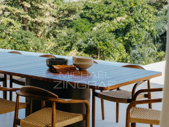 Table en bois avec chaises en rotin sur une terrasse avec vue sur la nature verdoyante.