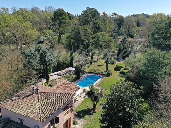 A spacious backyard with a rectangular blue pool, surrounded by trees and a stone patio, adjacent to a tiled-roof house.