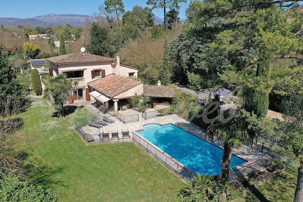 A Mediterranean-style house with tiled roof and a bright blue rectangular pool in a fenced yard, surrounded by trees and hills.