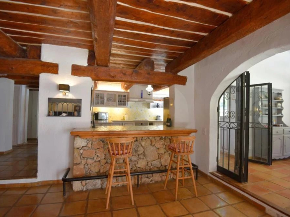Rustic kitchen with a stone-bar counter, two wooden bar stools, exposed wooden beams, and an arched doorway leading to a bright room with shelves.