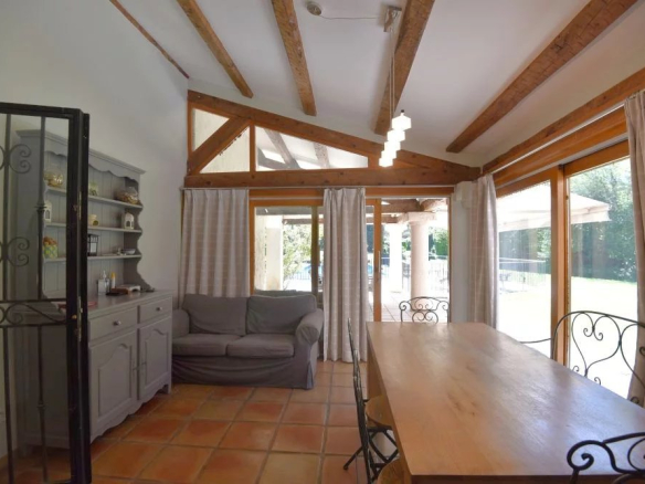 Sunlit dining area with a long wooden table, wrought-iron chairs, and a gray sofa near large glass doors.