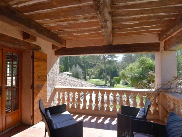 Covered terrace with wooden beams and wicker seating, overlooking a sunny garden with palm trees outside the balustrade.