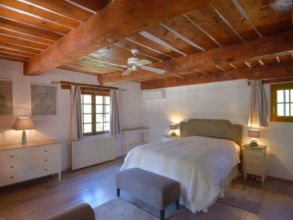 Rustic bedroom with exposed log beams, a king-size bed with a white quilt, two nightstands with lamps, and wooden floors atop a neutral rug.
