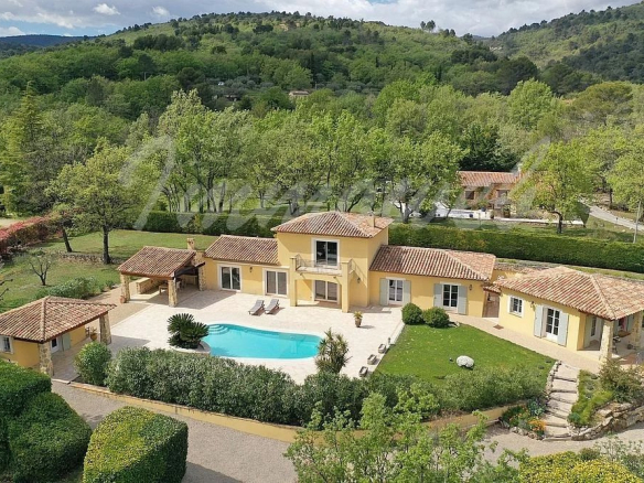 A yellow villa with terracotta roofs and a curved blue pool in a landscaped yard among trees (aerial view).