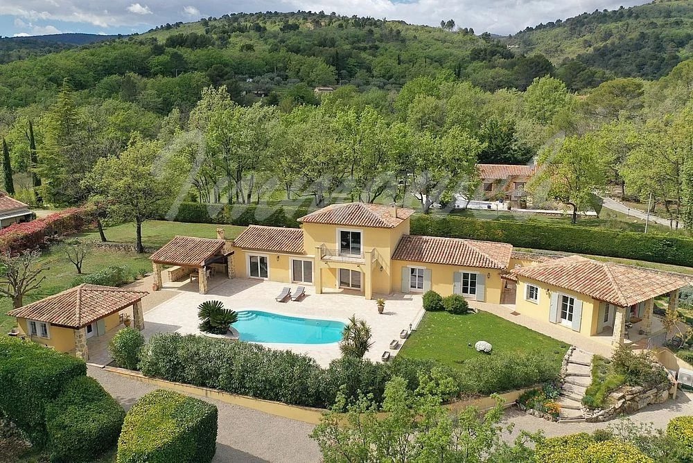 A yellow villa with terracotta roofs and a curved blue pool in a landscaped yard among trees (aerial view).