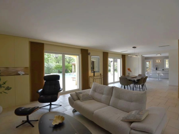 Open-plan living room with a beige sectional sofa, black lounge chair and ottoman, near large sliding doors to a garden and pool.