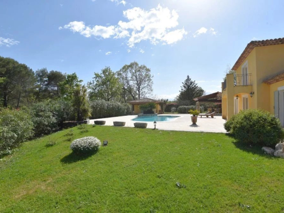 Sunny backyard with a rectangular pool, stone patio, and a yellow villa; lounge chairs on the patio and green lawn in the foreground.