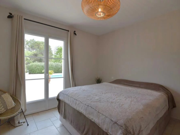 Bright bedroom with a queen bed, beige bedding, a wicker chair, and glass doors to a balcony outside.