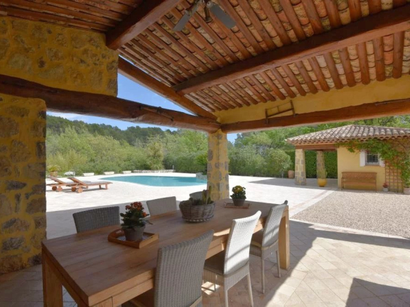 Outdoor covered patio with a wooden dining table and six wicker chairs overlooking a pool and garden area in bright daylight: warm stone columns and terracotta roof beams above.