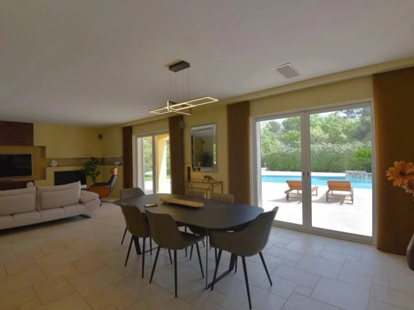 Open-plan living and dining room with a long dining table and gray chairs, sliding glass doors to a pool patio outside.