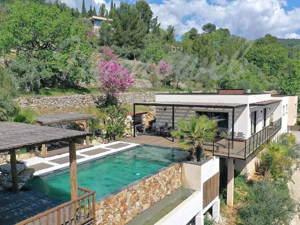 Contemporary hillside villa with a rectangular turquoise pool, stone retaining wall, and a shaded deck area surrounded by trees and flowering plants.