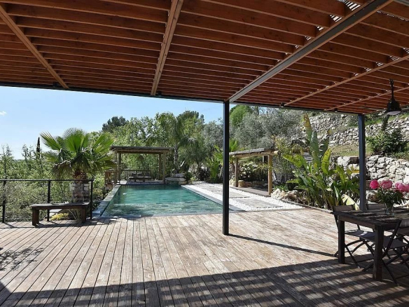 Open wooden deck with a rectangular pool, shaded patio area, potted plants, and a stone retaining wall on the hillside.