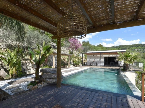 Outdoor pool area on a wooden deck under a thatched pergola, with tropical plants, a stone wall, and a hillside view.