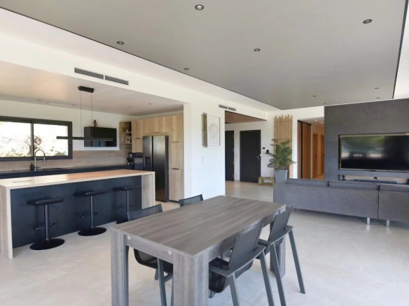 Open-plan living area with a modern kitchen island, black stools, and a dining table facing a gray sofa and wall-mounted TV.
