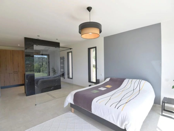 Modern bedroom with a gray accent wall, white bed with a striped runner, glass partition to an ensuite, a wooden wardrobe, and a potted plant.