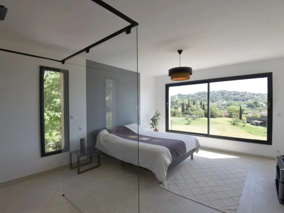 Bright minimalist bedroom with a large window overlooking hills; bed centered with white and gray bedding and a glass partition wall nearby.