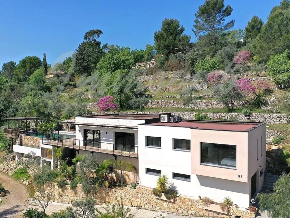 Modern white house on a terraced hillside with large windows and a red roof, surrounded by trees and blooming shrubs.