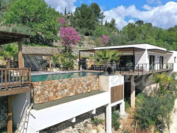 Modern hillside villa with a white facade, glass railings, and a stone-bordered infinity pool overlooking trees and hills on a sunny day.