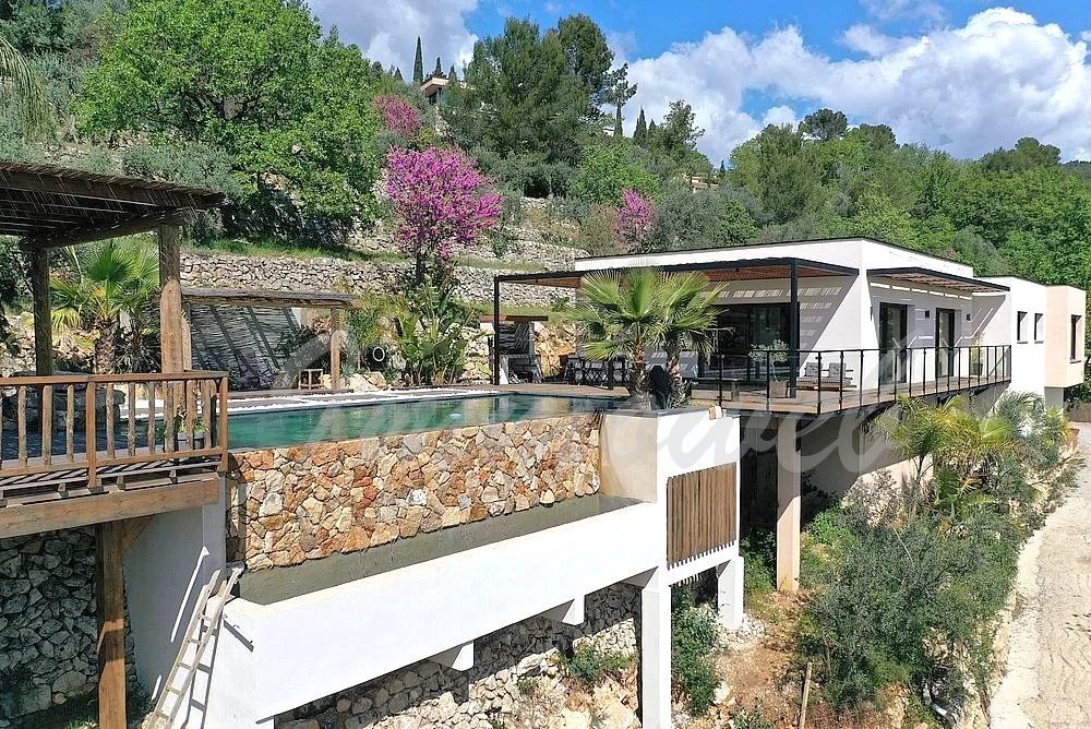 Modern hillside villa with a white facade, glass railings, and a stone-bordered infinity pool overlooking trees and hills on a sunny day.