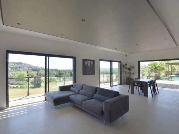 Open-plan living room with a gray sectional sofa and large sliding glass doors, showing a lawn and hills outside and a pool area beyond.